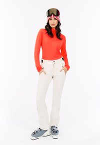 Felle oranje langmouwen top, witte skibroek met zakken, en grijze skischoenen met zwarte hardware. Witte achtergrond. Skibril op het hoofd.