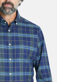 Camisa de cuadros azul con rayas verdes, marrones y azul claro. Cuenta con un cuello abotonado y botones blancos, fabricada en un tejido suave.