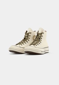 Zapatillas de deporte altas en lona crema con cordones verde oscuro, suela de goma, puntera texturizada y un parche de logo redondo en el lado exterior.
