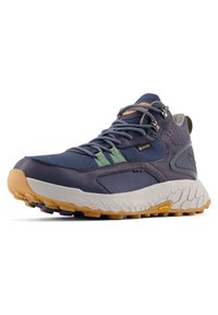 Zapato de senderismo Gore-Tex en tono azul marino y verde, con suela texturada blanca y suela Vibram color goma, ganchos laterales para los cordones y soporte acolchado para el tobillo.