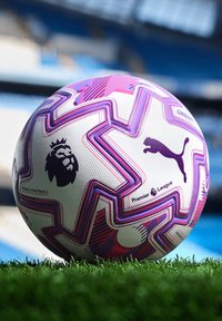 Pallone bianco della Premier League con disegni geometrici viola e rosa sull'erba, con uno sfondo sfocato di uno stadio.