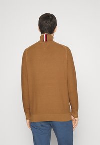 Homme de dos portant un pull marron texturé à col montant avec des rayures rouges, blanches et bleues, et un pantalon bleu.