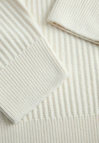 Tessuto a maglia a coste color crema con linee verticali e una consistenza morbida, caratterizzato da un dettaglio di risvolto per una maggiore dimensione.