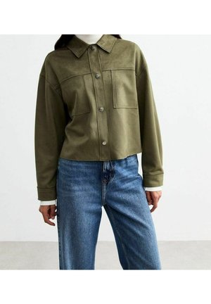 Kvinde iført en olivengrøn jakke med knapper og to brystlommer samt blå højtaljede jeans, stående mod en ensartet lys baggrund.