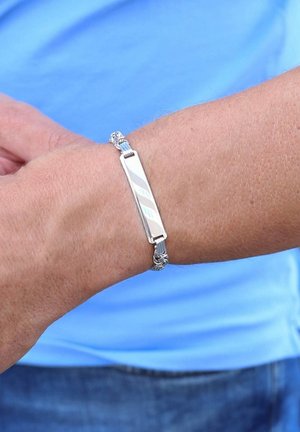 Silver bracelet with a sleek, oblong centerpiece and twisted metal accents, resting on a wrist against a light blue background.
