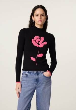 Vrouw draagt een zwarte ribgebreide trui met verhoogd roze bloemenmotief en manchetten, gecombineerd met lichtblauwe spijkerbroek, staand tegen een effen achtergrond.