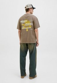Hnědé grafické tričko s žlutým textem na zádech, spárované s volnými gradientně zelenými džíny a tmavou kšiltovkou. Ležérní streetwear styl.