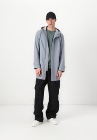 Lys grå vandtæt jakke med hætte og trykknaplukning, lagd over en grøn sweatshirt, parret med sorte cargo bukser og hvide sneakers.