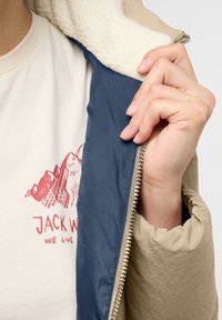 Beige jacka med blått innerfoder och vit fleecekrage, med en metallic dragkedja och en hand som håller i dragkedjan.