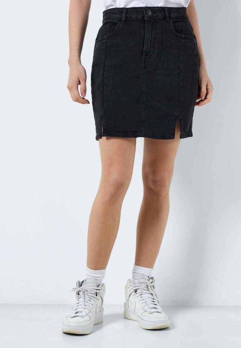 Personne portant une jupe en jean noire avec des fentes à l'avant, des baskets blanches, des chaussettes blanches et un haut blanc sur un fond clair uni.