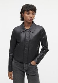 Schwarze Lederjacke mit Kragen, tailliertem Schnitt und Frontnahtverzierungen. Über dunkler Denim getragen, was eine elegante Textur zeigt.