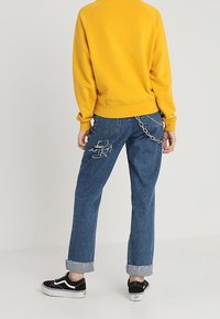 Sudadera amarilla, jeans azules desgastados con puños enrollados y zapatillas negras. Detalle de cadena notable en el bolsillo trasero.