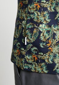 Marineblauw shirt met een groene en oranje tropische bladerenprint, gedragen met donkergrijze broeken, met zichtbare zijsplits en een klein merklabel.
