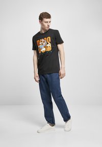 Svart bomulls t-skjorte med grafisk trykk som viser en tegneseriefigur og basketball; kombinert med blå jeans og hvite sneakers.