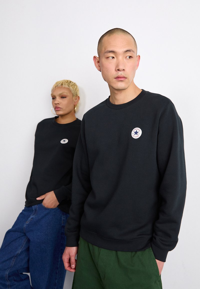 Sudadera negra de algodón, con cuello redondo y un parche de logo circular. Un modelo lleva vaqueros azules, el otro viste pantalones verdes.