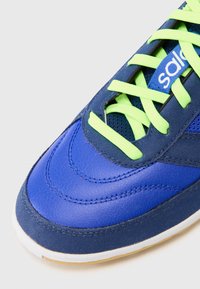 Blue athletic shoe featuring leather and mesh materials, with curvy stitching, neon green laces, and a white rubber sole.