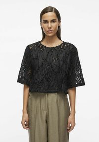 Top negro corto con tela texturizada y en capas, con acentos deshilachados; combinado con pantalones de pierna ancha en verde oliva claro.