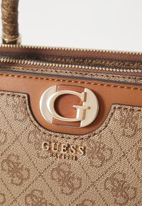 Brun mønstret håndtaske med glat tekstur, guldfarvet hardware og et fremtrædende logo. Lynlåslukning langs topkanten.