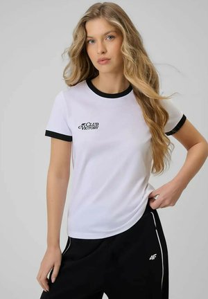 Młoda kobieta z długimi blond włosami, ubrana w białą koszulkę z czarnym wykończeniem i czarne spodnie z białym logo i paskami na szarym tle.