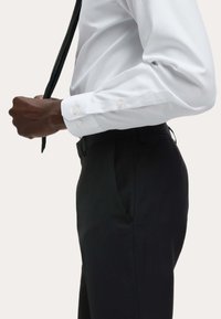 Biała koszula z długim rękawem na guziki z perłowymi guzikami, w połączeniu z czarnymi dopasowanymi spodniami, noszona z czarnym krawatem. Gładka faktura materiału.