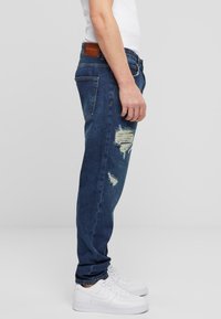 Jeans de mezclilla azul oscuro con un corte recto, con parches desgastados y una etiqueta en cuero marrón en la cintura, combinados con zapatillas blancas.