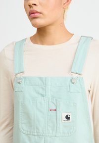 Svetlozelené bavlnené overaly s nastaviteľnými popruhmi, predným vreckom a štítkom s logom Carhartt. Obsahujú strieborné kovanie a minimalistický dizajn.