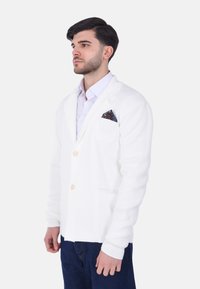Cardigan lavorato a maglia bianco con due bottoni, una tasca sul petto che presenta un fazzoletto colorato con motivo, e una vestibilità comoda. Dettaglio del tessuto testurizzato visibile.