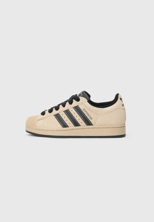 Beige semsket skinn-sneaker med svarte detaljer, med tre svarte striper, gummiforsterket tåhette og en flat svart såle. Snørene er svarte.