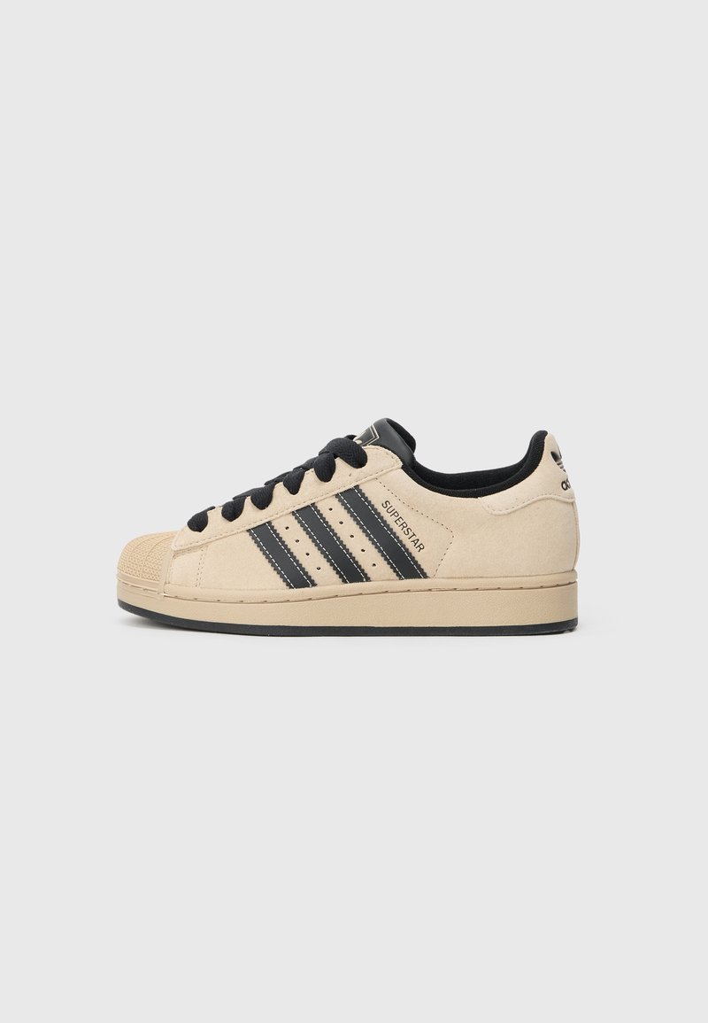 Μπεζ σουέντ αθλητικό παπούτσι με μαύρες λεπτομέρειες, που διαθέτει τρεις μαύρες γραμμές, λαστιχένια προστασία στα δάχτυλα και επίπεδη μαύρη σόλα. Τα κορδόνια είναι μαύρα.