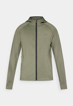 Veste vert olive avec une fermeture éclair intégrale à l'avant, une capuche et un accent de fermeture éclair noir. Présente des détails de coutures et un logo subtil sur la poitrine.