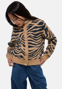 Jeune femme portant un cardigan beige et noir à motif animal avec des boutons, associé à un jean bleu ample, regardant vers le bas.