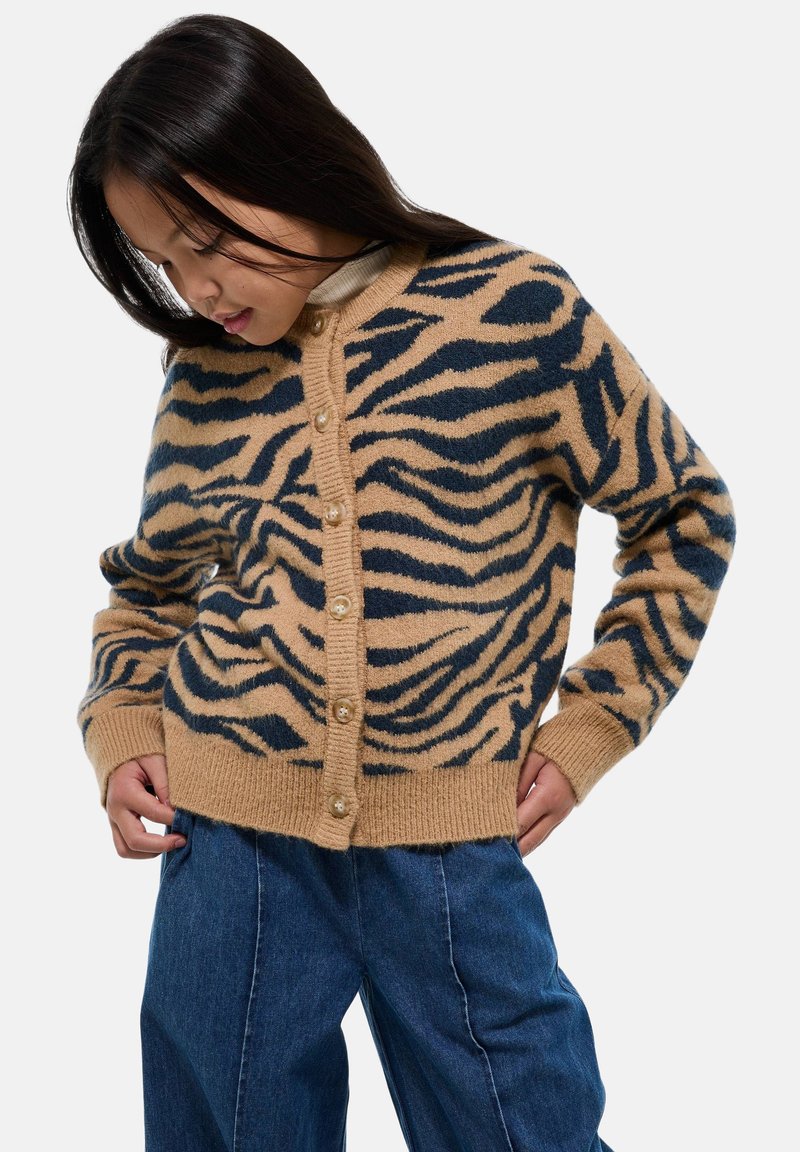Jeune femme portant un cardigan beige et noir à motif animal avec des boutons, associé à un jean bleu ample, regardant vers le bas.