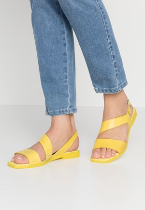 Voeten met blauwe jeans en felgele open sandalen met een bandje achter de hiel en lage vierkante hakken op een witte vloer.
