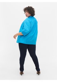 Modra bombažna bluzica s tremi četrtinami rokavov, nabranega videza na ramenih, oblečena z temno modrimi kavbojkami in črnimi čevlji s peto.