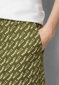 Pantalon vert olive avec un motif blanc répété d'épingles de sûreté et le nom de la marque « huf ». Confectionné dans un tissu résistant avec des poches latérales.