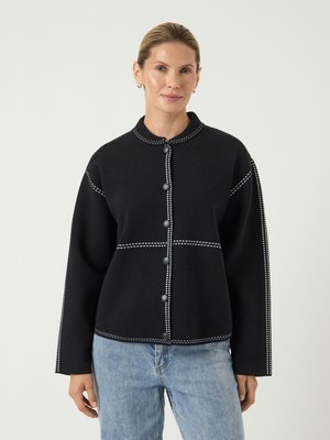 Svart knäppt jacka med rund krage, med vit sömnad, långa ärmar och stora grå knappar; kombinerad med ljusblå denimjeans.