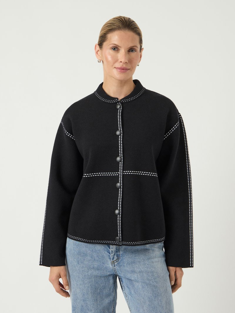 Μαύρο σακάκι με κουμπιά και στρογγυλέα γιακά, με λευκή ραφή, μακριά μανίκια και μεγάλα γκρι κουμπιά. Συνδυάζεται με ανοιχτό μπλε τζιν.