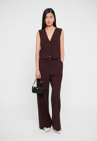 Burgundy sleeveless vest with buttons, paired with wide-leg trousers. Model holds a small black handbag. Simple, elegant silhouette.