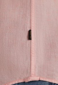 Tela de lino de color rosa claro con textura visible, con un borde cosido y una pequeña etiqueta negra en el lateral. Un dobladillo inferior curvado.