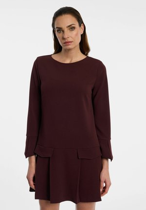 Woman with straight dark hair wearing a long-sleeve dark burgundy dress with front flap details standing against a plain light background.