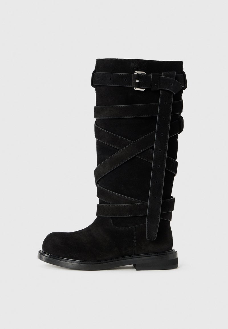 Cizmă din suede negru, până la genunchi, cu curele încrucișate și cataramă pe lateral. Talpa plată și vârful rotunjit subliniază designul structurat.