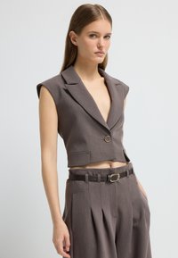 Zkrácený, bezrukávový blejzr z hnědé látky s hlubokým výstřihem ve tvaru V, uzavřením na jeden knoflík a detailním předním kapsou, skombinovaný s kalhotami s vysokým pasem.
