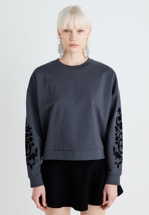 Grey sweatshirt with long sleeves, featuring black floral embroidery on the arms. Ribbed neckline and hem. Worn with a black skirt.
