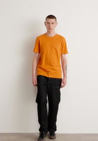 Orange kortærmet t-shirt med et subtilt logo på brystet, parret med mørkegrå cargo bukser og mørkt fodtøj, mod en neutral baggrund.
