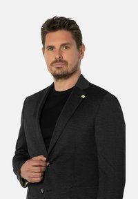 Man met kort bruin haar en baard die een donker geblokte blazer over een zwart shirt knoopt, recht vooruit kijkend tegen een effen achtergrond.