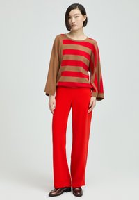Gestreifter braun-rot gestrickter Pullover mit weiten Ärmeln, kombiniert mit knallroten, weiten Hosen. Braune Schuhe runden das Outfit ab.