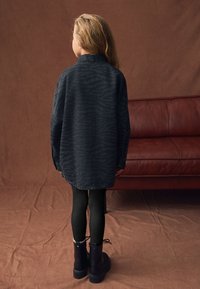 Maglietta nera con motivo a zebra, vestibilità oversize, maniche lunghe, dettagli con bottoni, abbinata a leggings neri e stivali neri robusti. Lo sfondo è marrone.