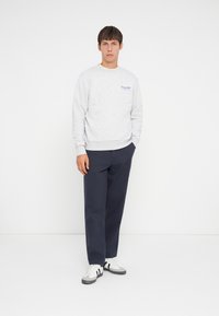 Light gray sweatshirt with ribbed cuffs and hem, featuring a blue logo. Paired with dark navy trousers and black-and-white sneakers.