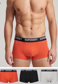 Bóxer naranja con una cinturilla elástica negra que presenta la marca "SUPERDRY". Mostrados con variantes en negro y gris debajo.