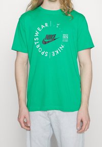 Vihreä puuvillapaita lyhyillä hihoilla, jossa on musta Nike-logo ja pyöreä tekstisuunnittelu. Istuvuus on rento ja kaula-aukko on standardikokoinen.